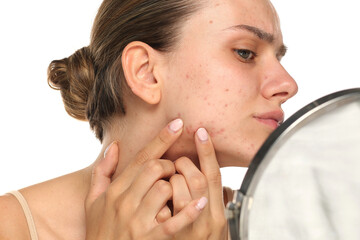 Studio shot of young woman examining acne on face in mirror on white background.