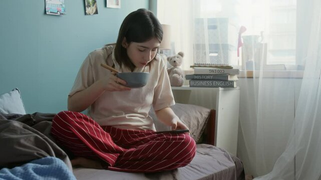Full length shot of teenage girl eating breakfast cereal while sitting in bed and using smartphone, gen Alpha