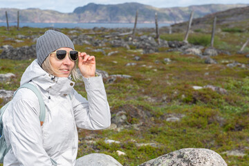 Woman in a white jacket adjusting her sunglasses while sitting among tundra rocks in Teriberka Nature Reserve with mountains and sea