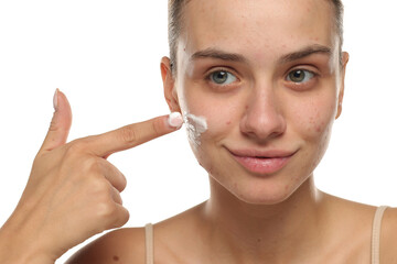 Young woman smiling while applying face cream on acne-prone skin, studio shot on white background