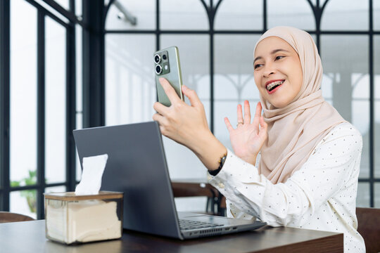 Asian Muslim woman wearing hijab sits at a table in a modern cafe using a laptop and smartphone for a video call.