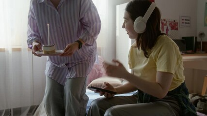 Caucasian girl using mobile phone while sitting on bed in dorm room and friend surprising her with Birthday cake - Powered by Adobe