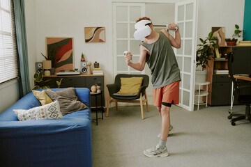Caucasian teenage boy wearing VR headset standing in living room exercising with motion controllers...