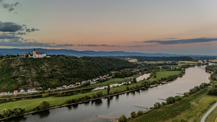 Panoramablick auf die Donau bei Sonnenuntergang, mit einer Kirche auf einem Berg und sanften Hügellandschaften