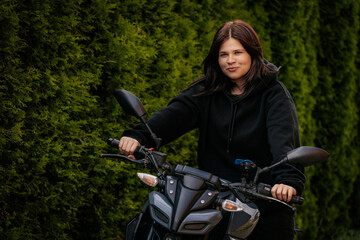 Young woman in black hoodie smiles while sitting on a motorcycle, set against a backdrop of lush...
