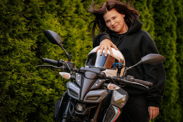 Young woman in black hoodie smiles while sitting on a motorcycle, set against a backdrop of lush green hedges on a sunny day.