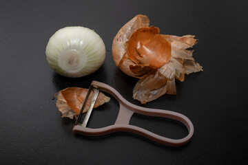 Peeled onion and onion peel next to it on black table and vegetable peeling knife.Close up of dry...