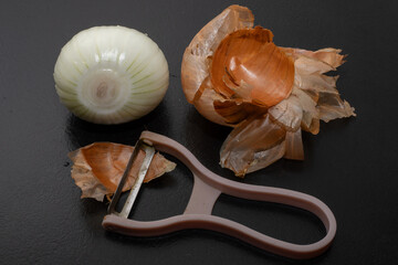 Peeled onion and onion peel next to it on black table and vegetable peeling knife.Close up of dry...