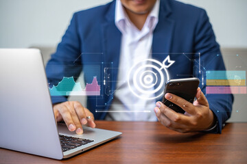 Strategic Business Growth A Professional Using a Smartphone and Laptop with a Holographic Target and Graph Overlay Representing Goals and Data Analysis
