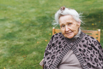 Smiling elderly woman 80-90 years with grey hair sitting on a wooden chair outdoors, wearing warm...