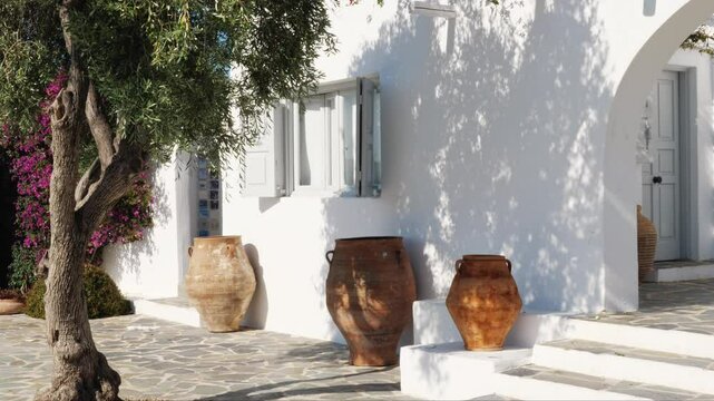 Large vintage terracotta pots near a whitewashed Greek house with open shutters, stone pavement, olive tree shadow, and pink flowers. Shot in Athens, Greece. Sunny day. No people.
