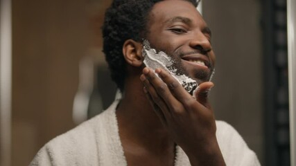 African American man shaving beard applying foamy gel in bathroom bearded guy smear foam shave procedure looking at mirror unshaven bristle male touching skin prepare face care hygiene cosmetic beauty - Powered by Adobe
