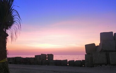 A serene sunset over a calm sea is framed by a palm tree to the left and a wall of concrete blocks...