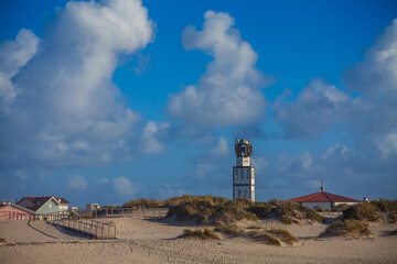 A tall, white clock tower with a distinct top stands prominently amidst sandy dunes