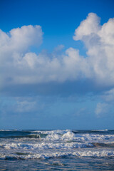 A dynamic seascape captures foamy waves breaking on a sandy shore under a brilliant blue sky dotted with white clouds
