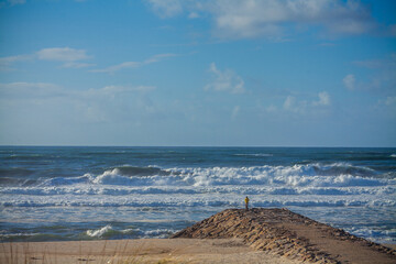 A person stands alone at the end of a rocky breakwater, observing the vast ocean