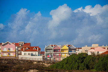 A line of vibrant, colorful striped houses with red-tiled roofs stands behind a wooden boardwalk and green vegetation