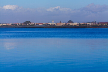 A serene blue lagoon reflects a distant coastal town with red-roofed buildings