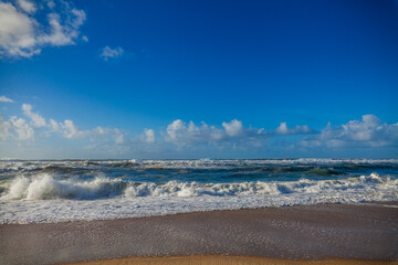 A dynamic seascape captures foamy waves breaking on a sandy shore under a brilliant blue sky dotted with white clouds