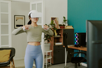 Teenage girl standing in modern room using VR headset and controllers, engaging in virtual reality...