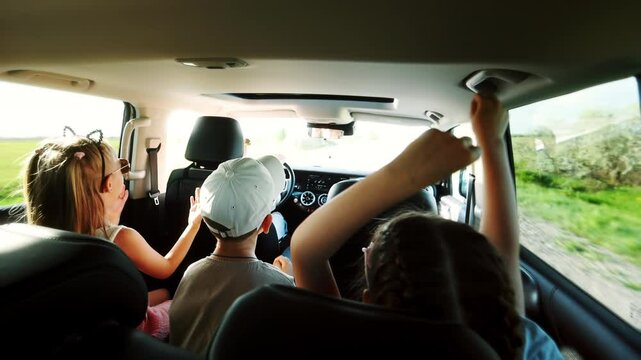 Mother takes her children by car to nature on a sunny summer day along a country road. The children are having fun in the back seat, singing and raising their hands, dancing to the music