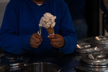 A hand in a blue hoodie scoops velvety ice cream into a crispy waffle cone inside a modern parlor, capturing the inviting swirl and mouthwatering detail