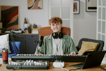 Caucasian teenage boy sitting at desk recording music, wearing headphones around neck, playing...