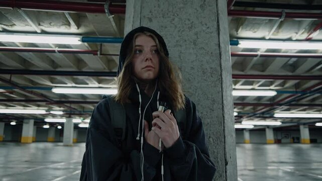 Young woman standing in deserted concrete parking garage, wearing hoodie and backpack, vaping with contemplative expression while exhaling vapor cloud against shadowy urban background