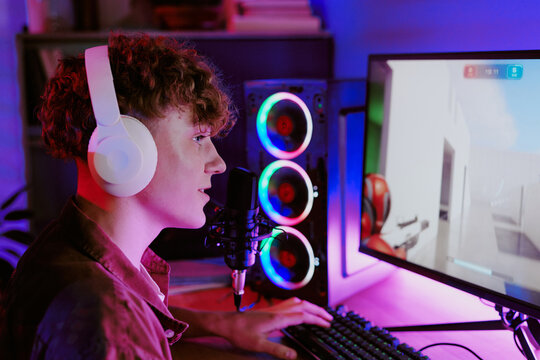 Caucasian teenage boy wearing headphones sitting at desk playing video game on computer while live streaming and speaking into microphone 