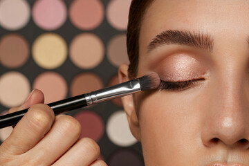 A close-up beauty shot of eye makeup application. A hand holding a black makeup brush applies eyeshadow to a closed eye. The brush has a soft, tapered tip and is made of synthetic bristles.