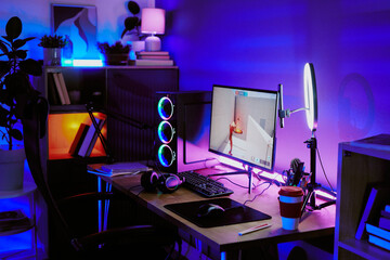 Modern home gaming setup featuring desktop computer with RGB lighting, large monitor displaying video game, professional microphone, headphones, VR headset, coffee cup on desk