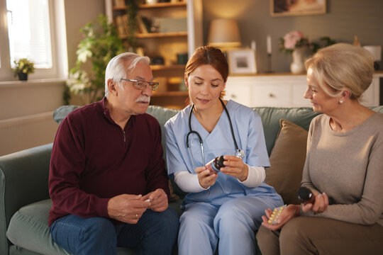 In a cozy living room, a female home health care worker wearing scrubs explains medication dosage and instructions using a pill bottle to an elderly couple sitting on a sofa.
