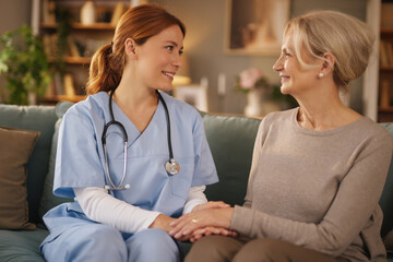 In a home setting, a friendly nurse in scrubs holds hands and smiles at an older woman. The pair is...
