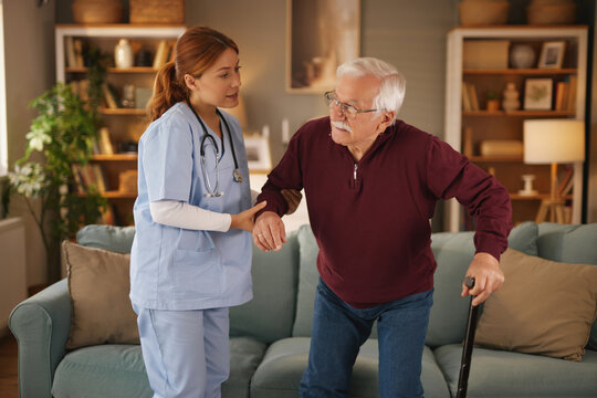 A female home care nurse in scrubs assists an elderly man using a cane to stand up from his couch. The nurse holds his arm while the man uses his cane for support. - Powered by Adobe