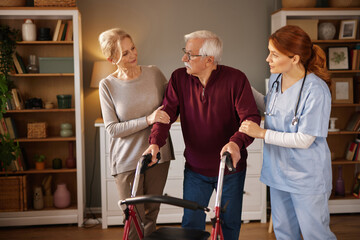 An elderly man uses a walker for mobility in a living room, with his wife and a home care nurse assisting him. It is daytime and the scene takes place in a cozy home setting.