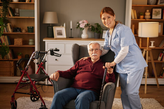 A female home care nurse in scrubs comforts an elderly man seated in a chair inside his house. The nurse has her hand on his shoulder as the man holds a cane, with a walker beside him.