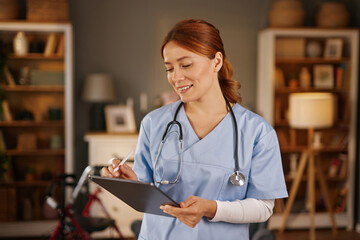 A female healthcare worker, wearing light blue scrubs and a stethoscope, uses a stylus to take notes on a tablet. She is in a living room with bookshelves and warm lighting.