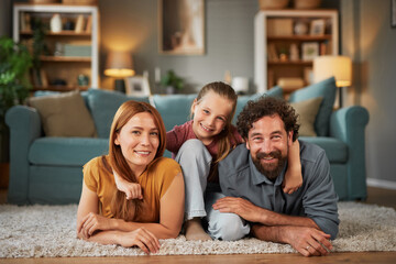 A mother, father, and daughter are lying on the carpet of a warm living room, smiling and embracing...