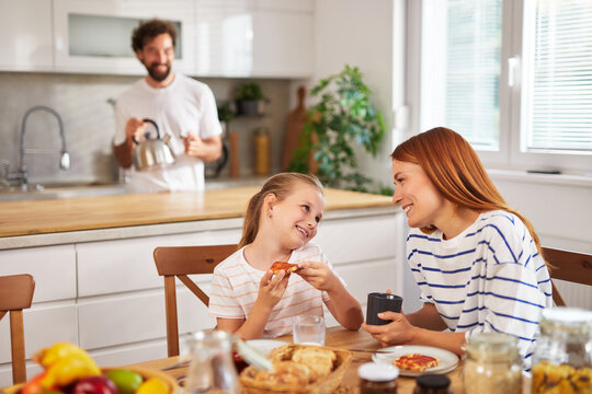 A young girl and her mother share a joyful moment over breakfast, smiling and talking, while a dad prepares tea in the background. The kitchen is bright and inviting.