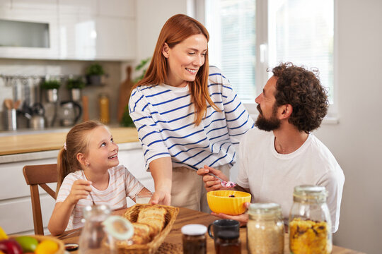 A cheerful family shares a joyful breakfast moment in their bright kitchen. The woman serves food while the girl and man engage in happy conversation, creating warm memories.