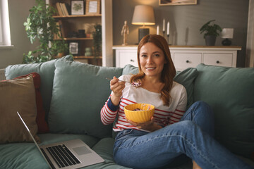 A woman relaxes on a comfortable sofa, holding a bowl of cereal with a spoon while her laptop is...