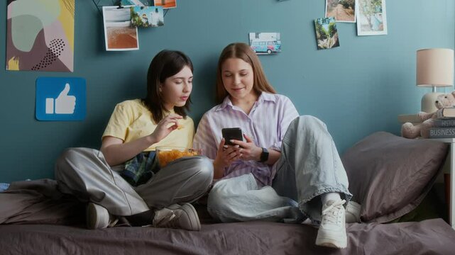Full length front view shot of two teenage girls sitting on bed in dorm room and looking at smartphone screen together while eating snacks