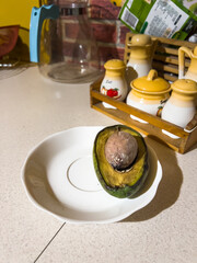 Sliced, rotten avocado with a pit on a white plate, lying on the kitchen counter, in the background you can see spices and a glass jug, in natural light