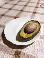 Rotten avocado with a large pit, lying on a white plate on a patterned tablecloth, showing the waste of food and staleness
