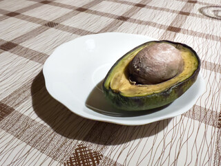 A single half of a rotten avocado with a large pit, placed on a white plate, resting on a brown and beige grid tablecloth