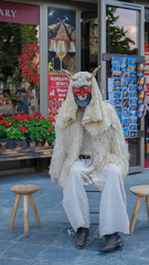 Man dressed in traditional Hungarian Busó costume with a horned mask and wool cloak, sitting outside a souvenir shop in Budapest.