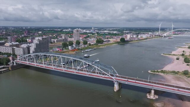 Sweeping aerial pan of Nijmegen, Netherlands, showcasing the Waal River, De Oversteek bridge, railway bridge, sandy banks, and wind turbines.