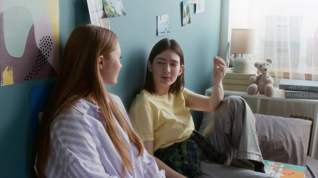 Medium shot of two smiling teenage girls chatting sitting on bed in dorm room, friends or roommates interacting