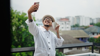 Happy asian man drinking coffee and taking a selfie on his balcony