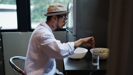 Asian man wearing a hat and glasses enjoying a healthy salad in his modern kitchen